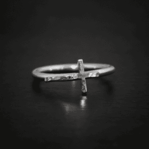 Close up of the "Creator" Ring. Made out of sterling silver. Ring has a thin, hammered oxidized cross going sideways across the ring band so that the top of the cross is on the right side of the ring band, the bottom of the cross is on the left side, and the two sides of the cross are sticking off of the ring band from the center of the cross. Ring is sitting against a dark background.